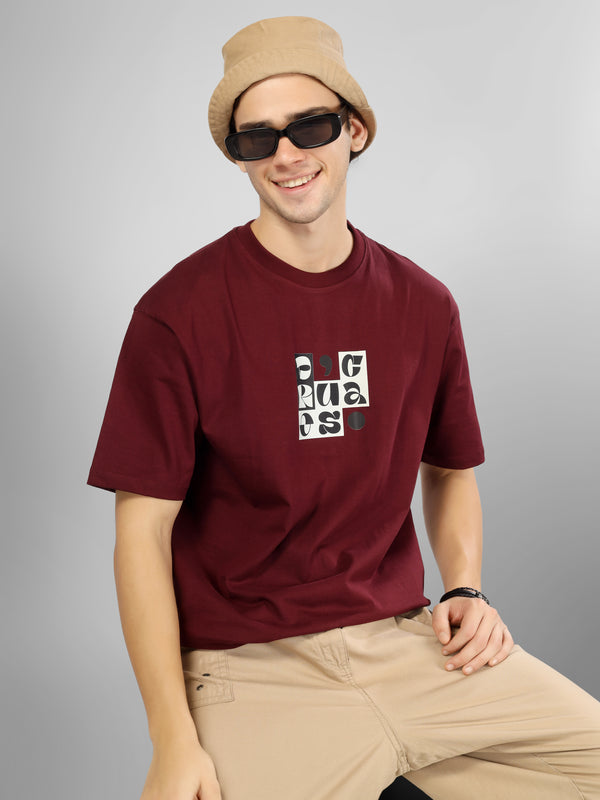 Male model wearing a maroon dcavros T-shirt featuring a black-and-white abstract center logo print, paired with beige pants.