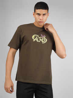 Male model wearing olive green dcavros unisex T-shirt with a yellow and pink "Cavros" bull logo on the chest, styled with black pants.
