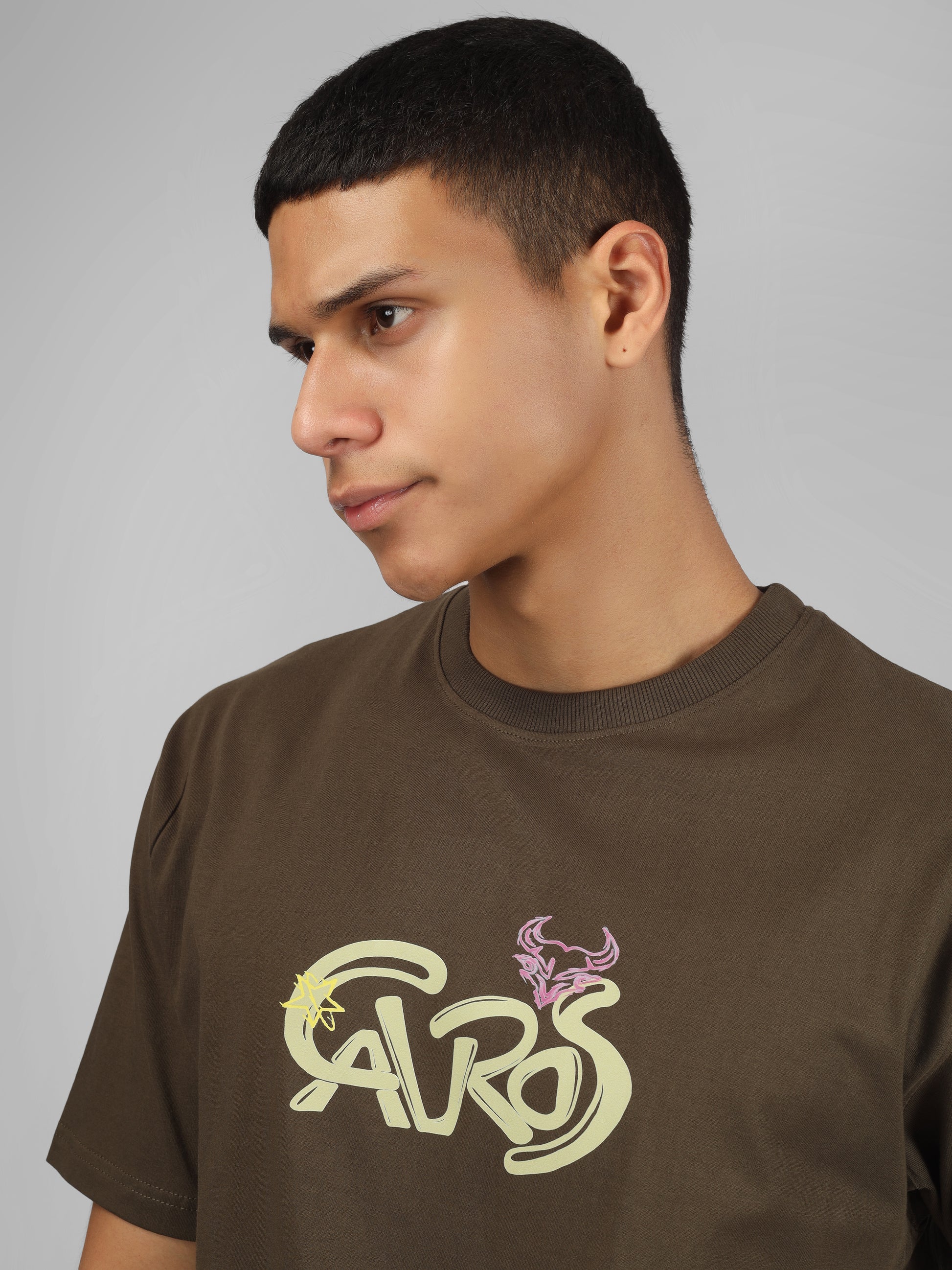 Male model wearing olive green dcavros unisex T-shirt with a yellow and pink "Cavros" bull logo on the chest, styled with black pants.