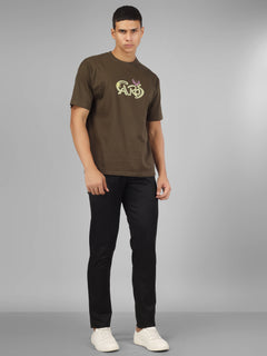 Male model wearing olive green dcavros unisex T-shirt with a yellow and pink "Cavros" bull logo on the chest, styled with black pants.