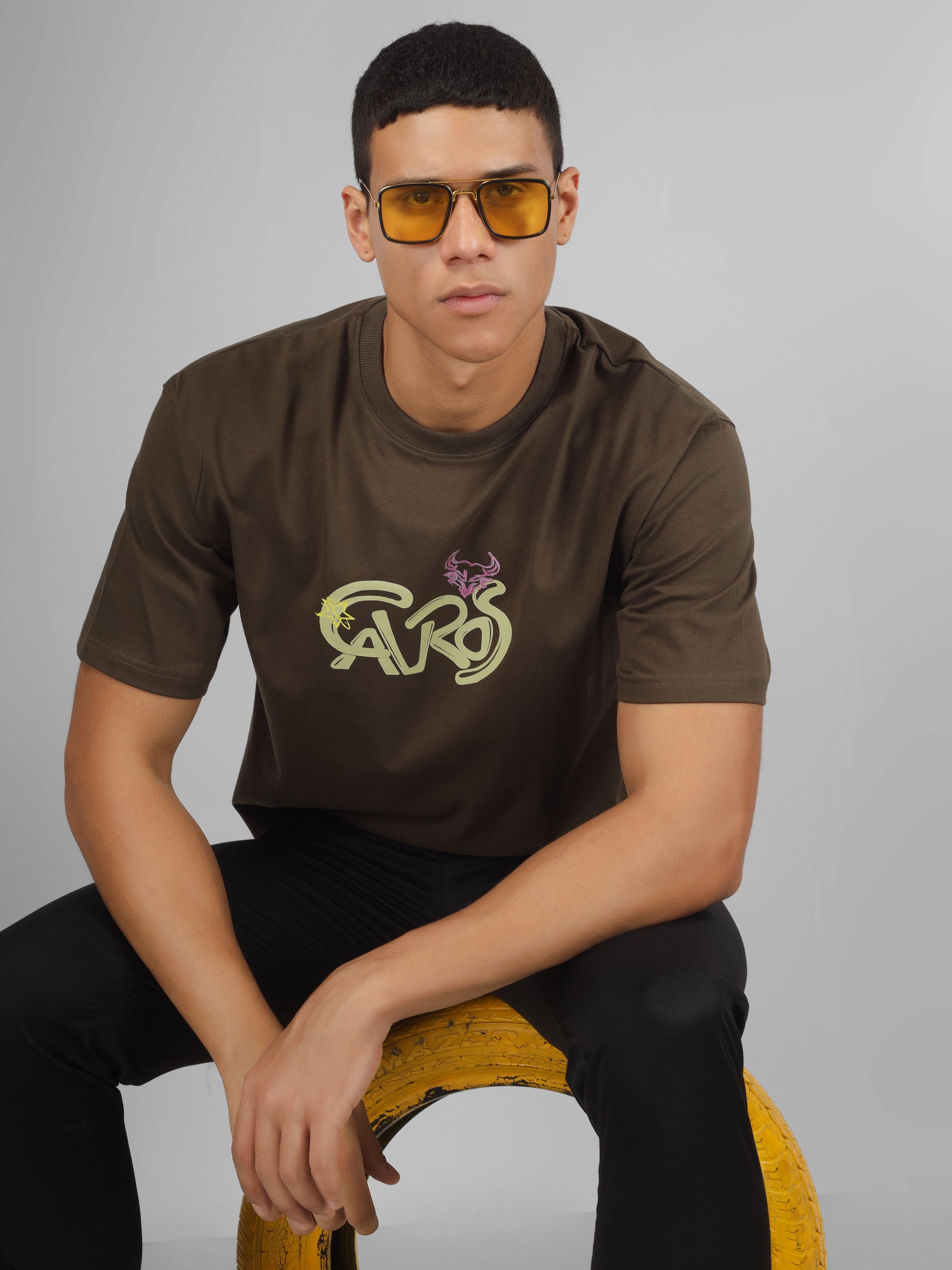 Male model wearing olive green dcavros unisex T-shirt with a yellow and pink "Cavros" bull logo on the chest, styled with black pants.