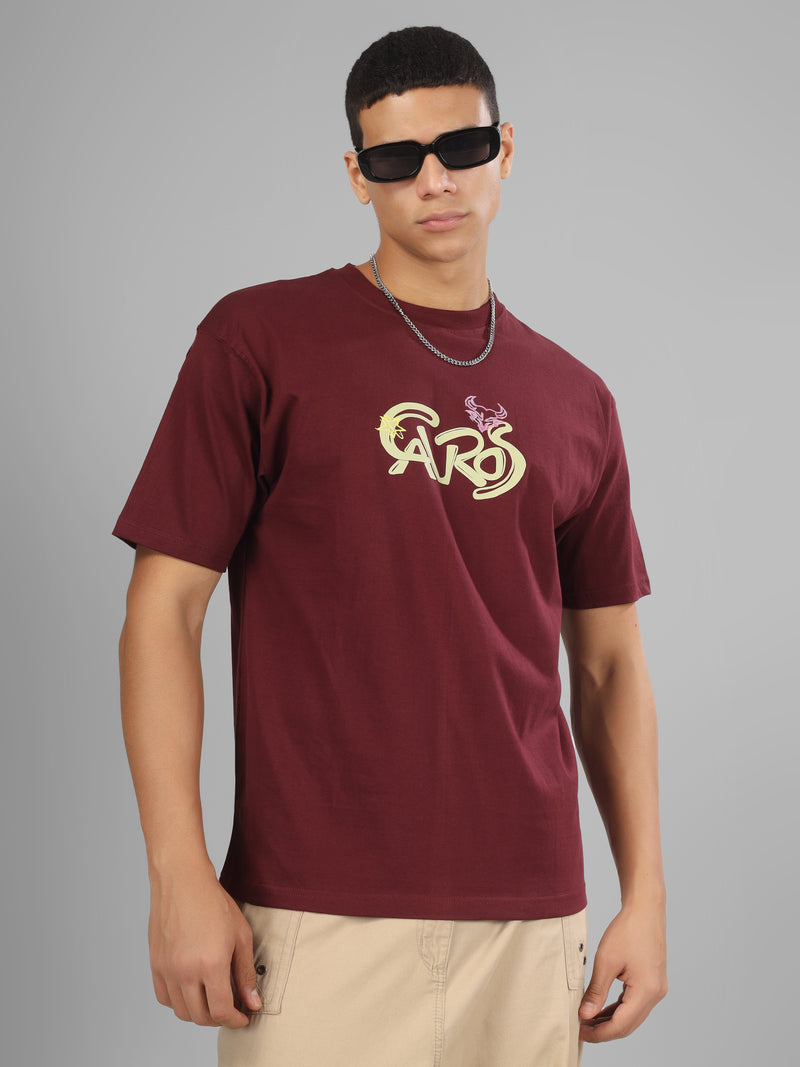 Male model wearing a deep maroon dcavros unisex T-shirt with a stylized yellow and pink "Cavros" bull logo on the front, paired with beige pants.