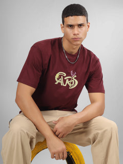 Male model wearing a deep maroon dcavros unisex T-shirt with a stylized yellow and pink "Cavros" bull logo on the front, paired with beige pants.